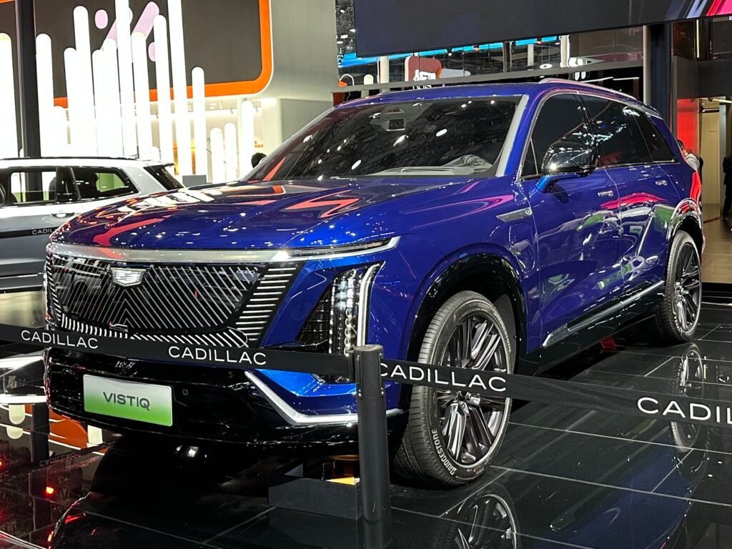 A blue Cadillac VISTIQ, a three-row electric SUV, on an auto-show stand, viewed from the front three-quarter angle.