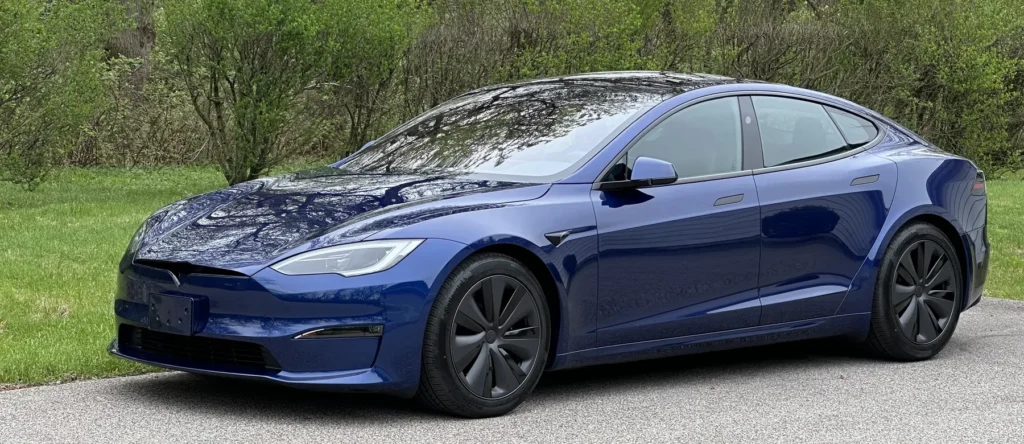 Blue 2024 Tesla Model S parked beside a wooded road, viewed from the front three-quarter angle.